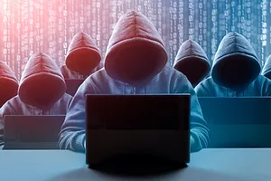 SEVEN HOODED FIGURES, WHOSE FACES CANNOT BE SEEN, ARE SITTING IN FRONT OF LAPTOPS WITH DIGITAL BINARY IN THE BACKGROUND.