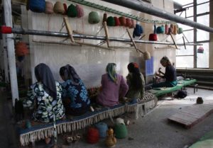 FIVE WOMEN WEARING HIJABS CAN BE SEEN SITTING ON POORLY-MADE BENCHES WORKING IN A DIMLY-LIT CONCRETE ROOM. 