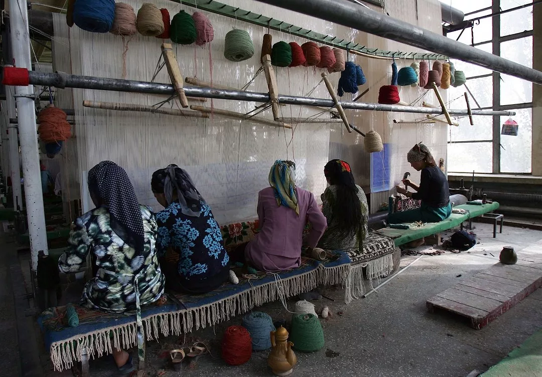 FIVE WOMEN WEARING HIJABS CAN BE SEEN SITTING ON POORLY-MADE BENCHES WORKING IN A DIMLY-LIT CONCRETE ROOM.