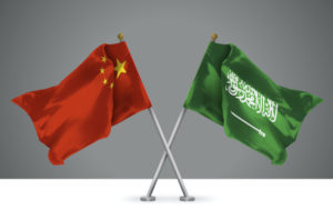 3D ILLUSTRATIONS OF THE WAVING CROSSED FLAGS OF CHINA AND THE KINGDOM OF SAUDI ARABIA SIT ON TOP OF A WHITE DESK WITH A GREY BACKGROUND.