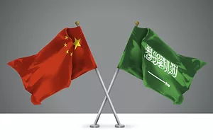 3D ILLUSTRATIONS OF THE WAVING CROSSED FLAGS OF CHINA AND THE KINGDOM OF SAUDI ARABIA SIT ON TOP OF A WHITE DESK WITH A GREY BACKGROUND.