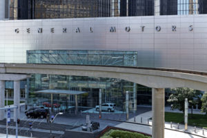 A SILVER BUILDING CAN BE SEEN WITH A LARGE GLASS ENTRYWAY THAT HAS THE WORDS "GENERAL MOTORS" ABOVE IT. PEOPLE CAN BE SEEN LEAVING THE BUILDING. THERE ARE FOUR GM CARS IN A ROUNDABOUT LEADING TO THE BUILDING ENTRANCE.