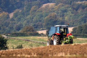 AN AGRICULTURAL TRACTOR CAN BE SEEN PLOUGHING A BROWN AND GREEN FIELD IN FRONT OF A ROLLING HILLSIDE FILLED WITH TREES.