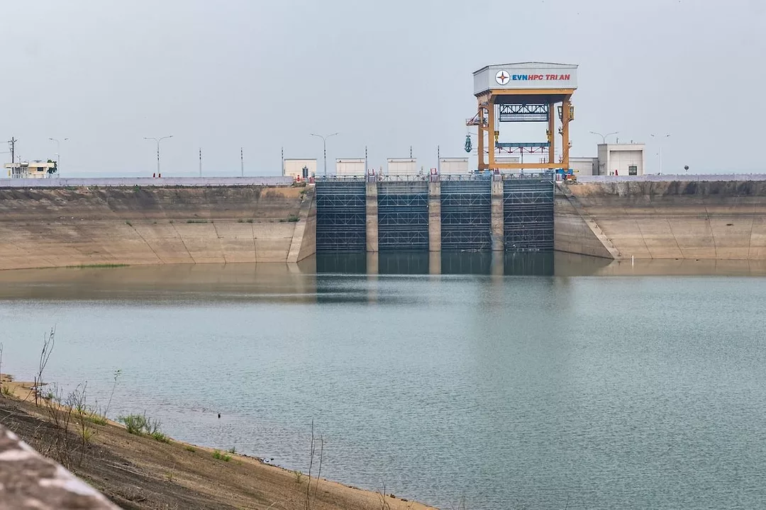 A BODY OF WATER LEADS TO THE OUTSIDE OF A HYDROPOWER PLANT WITH A TOWER CONNECTED THAT HAS THE WORDS "EVN" IN BLUE FOLLOWED BY "HPC TRI AN" IN RED