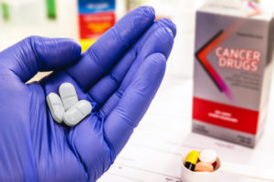 A HAND WEARING A BLUE GLOVE CAN BE SEEN HOLDING THREE WHITE PILLS WITH A SMALL CONTAINER OF VARIOUS MEDICATIONS BELOW IT. IN THE BACKGROUND, A GREY AND RED BOX IS LABLED "CANCER DRUGS."