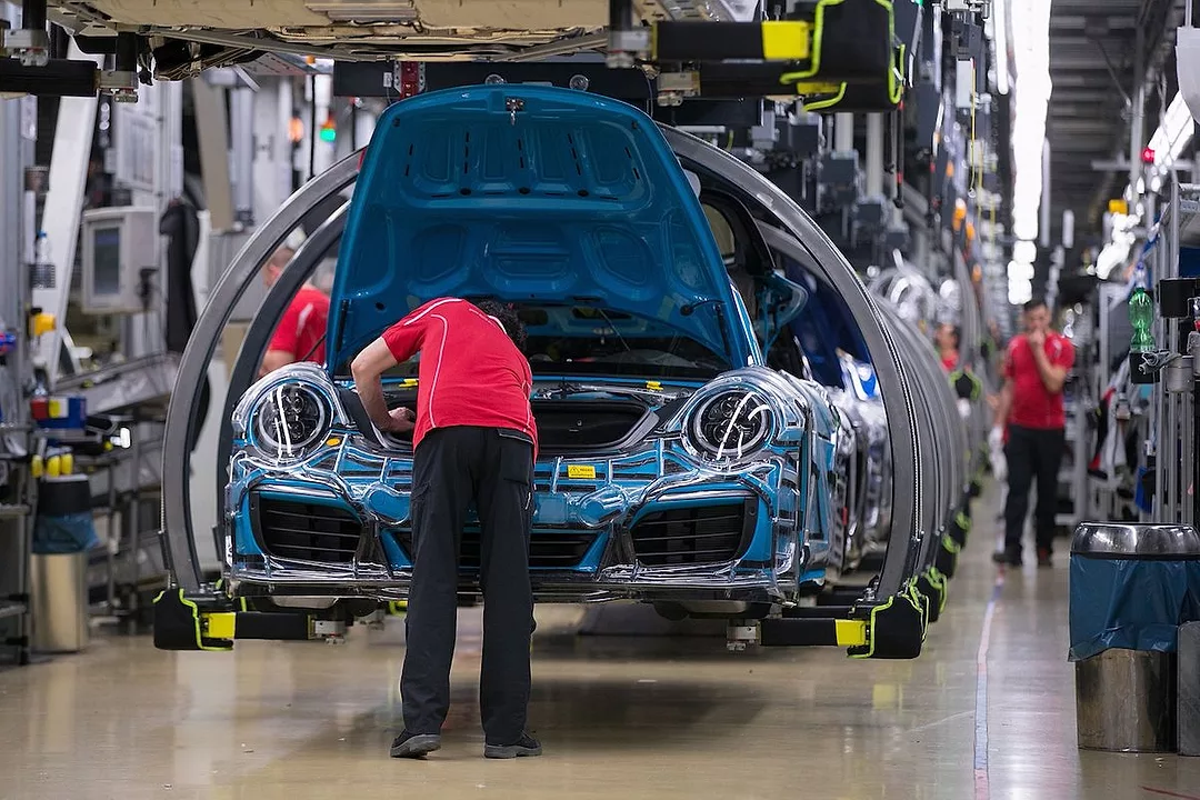 A PERSON WORKING IN A FACTORY WEARING A RED SHIRT AND BLACK PANTS IS BENT DOWN LOOKING UNDER THE OPENED HOOD OF A PARTIALLY-ASSEMBLED PORSCHE.
