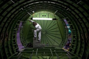 A PERSON IN A WHITE JUMPSUIT IS WORKING INSIDE A BOEING 737. THE GREEN WALLS OF THE AIRCRAFT ARE ILLUMINATED BY A SMALL FLORESCENT LIGHT ABOVE THE WORKER.
