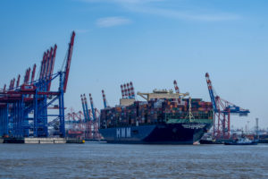 A LARGE CONTAINER SHIP WITH SEVERAL CONTAINERS IS LEAVING A PORT SURROUNDED BY CRANES. "HMM" IS WRITTEN ON THE SIDE OF THE CARRIER.