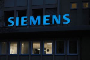 AN ILLUMINATED SIEMENS LOGO CAN BE SEEN AT NIGHT. BELOW IT ARE OPEN SHADES THAT REVEAL A DIMLY-LIT OFFICE.