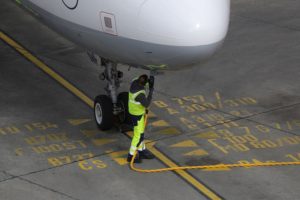 A GROUND CREW WORKER IN NEON YELLOW ATTIRE CAN BE SEEN CONNECTING A FUEL PUMP TO THE UNDERSIDE OF AN AIRPLANE.