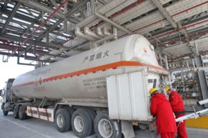 TWO PEOPLE IN RED JUMPSUITS AND YELLOW HELMETS CAN BE SEEN WORKING ON THE BACK OF A LNG TRUCK THAT HAS CHINESE WRITING ON THE SIDE OF IT.