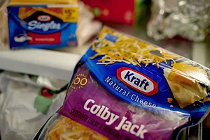 A BLUE BAG OF KRAFT COLBY JACK SHREDDED CHEESE CAN BE SEEN BEING HELD IN FRONT OF AN OPEN REFRIGERATOR WITH KRAFT CHEESE SINGLES AND OTHER GROCERIES BEHIND IT IN THE BACKGROUND.