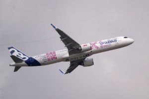 A WHITE AIRBUS PLANE WITH BLUE AND PINK MARKINGS CAN BE SEEN FLYING IN A GREY SKY DURING THE PARIS AIR SHOW.