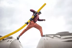 A PERSON IN AN ORANGE SUIT AND WHITE HELMET CAN BE SEEN STEPPING FROM THE TOP OF ONE TANK TRUCK TO ANOTHER WITH A YELLOW HOSE IN THEIR RIGHT ARM.