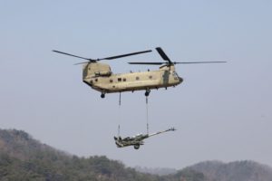 AN ARMY HELICOPTER CAN BE SEEN CARRYING A MILITARY WEAPON WITH MOUNTAINS IN THE BACKGROUND.