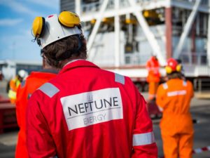 SEVERAL PEOPLE IN RED AND ORANGE JUMPSUITS APPEAR TO BE ON A SHIP. A PERSON IN A RED SUIT AND WHITE HELMET WITH A NEPTUNE ENERGY LOGO ON THEIR BACK CAN BE SEEN AT THE FOREFRONT OF THE PICTURE.