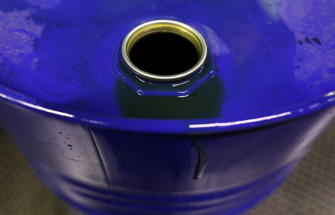 THE TOP OF A BLUE BARREL OF OIL CAN BE SEEN WITH THE CAP UNDONE. SOME SPOTS OF OIL ARE SPLATTERED ON THE BARREL.