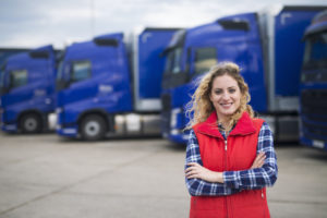 A WOMAN WITH DIRTY-BLONDE HAIR, A PLAID BLUE LONG-SLEEVE SHIRT AND A RED VEST CAN BE SEEN SMILING AND STANDING IN FRONT OF A LINEUP OF BLUE TRUCKS.