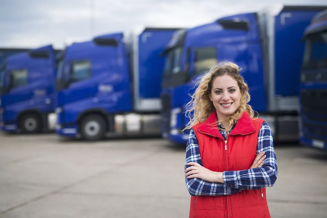 A WOMAN WITH DIRTY-BLONDE HAIR, A PLAID BLUE LONG-SLEEVE SHIRT AND A RED VEST CAN BE SEEN SMILING AND STANDING IN FRONT OF A LINEUP OF BLUE TRUCKS.