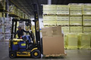 A PERSON OPERATING A DEWALT FORKLIFT IN A WAREHOUSE CAN BE SEEN WITH A PALLET OF BOXES ON THE LIFT PART OF THE MACHINE. 
