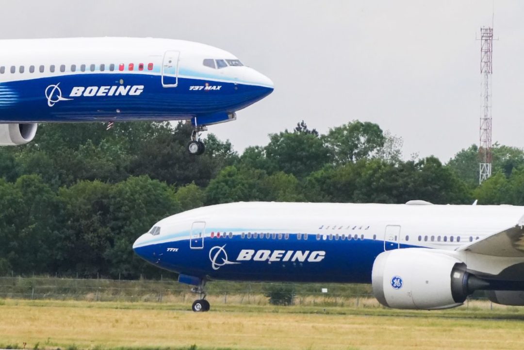 A WHITE AND BLUE BOEING 737 MAX AIRCRAFT IS LANDING BESIDE A WHITE AND BLUE BOEING B777-9 JET ON AN AIRPORT RUNWAY.