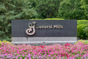 A MARBLE GENERAL MILLS SIGN SITS BEHIND A PATCH OF PINK FLOWERS. THE LETTERS SPELLING OUT GENERAL MILLS APPEAR TO BE MADE OF SILVER. 