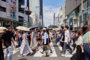 MANY PEOPLE CAN BE SEEN CROSSING THE STREET OF A BUSY SHOPPING DISTRICT IN JAPAN. A FEW PEOPLE ARE HOLDING UMBRELLAS.