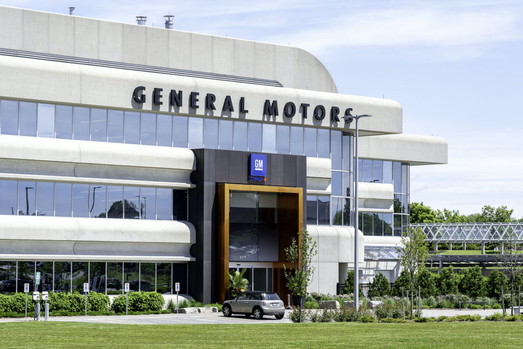 THE EXTERIOR OF A GM TECHNICAL CENTER CAMPUS CAN BE SEEN DURING A SUNNY DAY. A CAR IS PARKED RIGHT NEAR THE FACILITY'S FRONT ENTRANCE. 