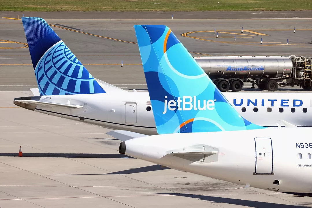 THE TAIL FINS OF TWO AEROPLANES, ONE JET BLUE, THE OTHER UNITED, SIT ON AN AIRPORT TARMAC.