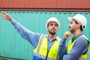 TWO WORKERS IN HI-VIS VESTS GESTURE IN FRONT OF A STACK OF SHIPPING CONTAINERS