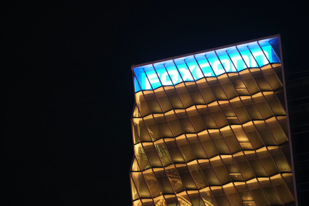THE BLUE AND WHITE FOXCONN LOGO IS DISPLAYED AT THE TOP OF A GLASS BUILDING AT NIGHT.