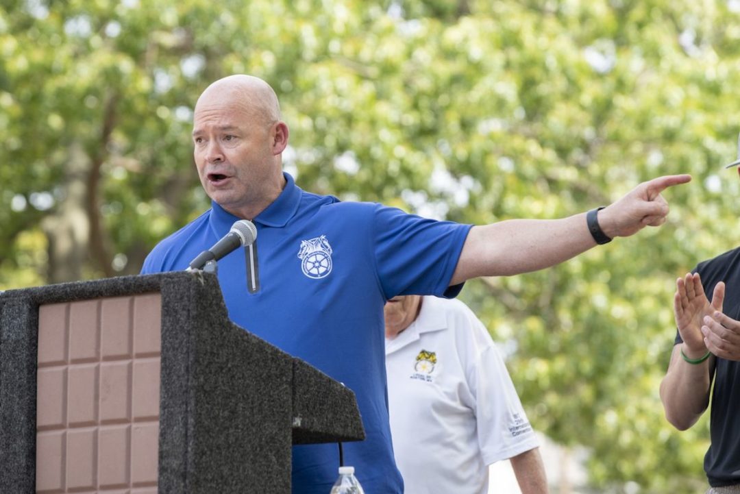 SEAN O'BRIEN, THE GENERAL PRESIDENT OF THE INTERNATIONAL BROTHERHOOD OF TEAMSTERS, IS IN A BLUE SHIRT SPEAKING BEHIND A PODIUM ON A STAGE AND POINTING LEFT WITH HIS HAND. TWO OTHER PEOPLE APPEAR TO BE ON THE STAGE.