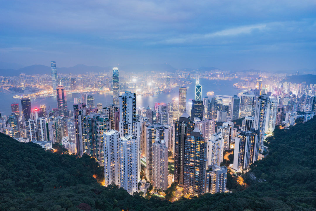 A DOWNWARD LOOKING VIEW OF DOWNTOWN HONG KONG CAN BE SEEN AT DUSK. SOME TREES ARE IN THE FOREGROUND OF THE IMAGE.