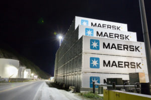 SEVERAL WHITE STORAGE CONTAINERS WITH THE MAERSK LOGO ON THEM ARE STACKED ON TOP OF EACH OTHER AT NIGHT NEXT TO A ROAD.