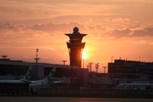 AN AIR TRAFFIC CONTROL TOWER SURROUNDED BY PLANES AT AN AIRPORT PARTIALLY BLOCKS OUT A SETTING SUN.