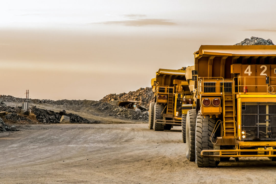 TWO YELLOW DUMP TRUCKS CARRYING MINERALS APPEAR TO BE DRIVING AWAY FROM A MINE.