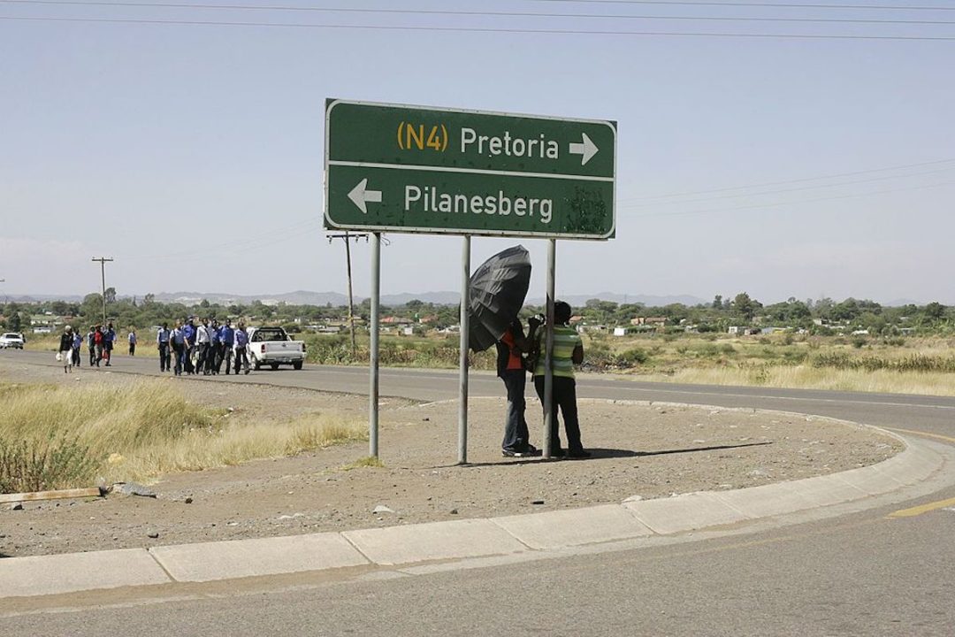 TWO PEOPLE CAN BE SEEN STANDING UNDERNEATH A ROAD SIGN THAT READS "(N4) PRETORIA" FOLLOWED BY "PILANESBERG." ONE OF THE PEOPLE IS HOLDING AN UMBRELLA.