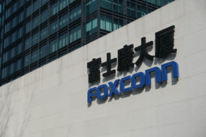 A WALL WITH THE FOXCONN LOGO ON IT CAN BE SEEN IN FRONT OF A LARGE GLASS BUILDING. THIS IS THE FOXCONN HEADQUARTERS IN SHANGHAI, CHINA.