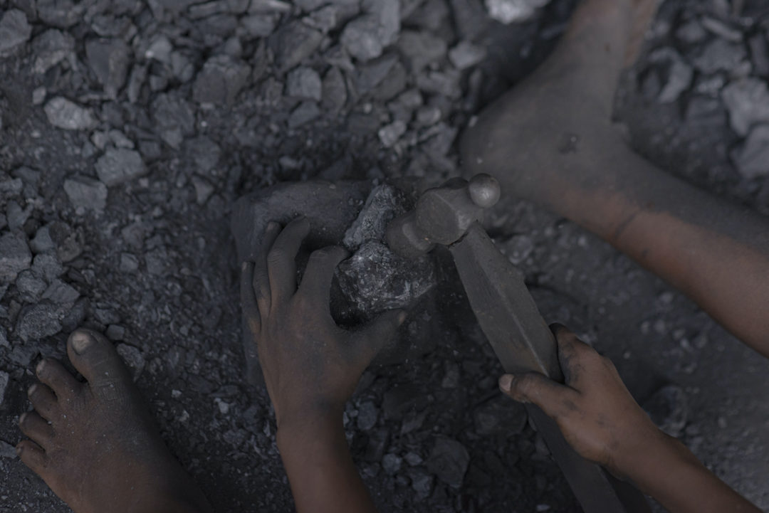 A SMALL PAIR OF HANDS IS HOLDING AN IRON HAMMER AND A PIECE OF COAL. A SMALL FOOT CAN BE SEEN BESIDE EACH HAND.