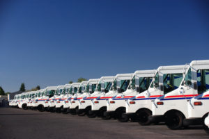 A FLEET OF UNITED STATES POSTAL SERVICE TRUCKS ARE PARKED NEXT TO EACH OTHER IN A PARKING LOT BELOW A CLEAR BLUE SKY.
