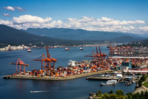 THE PORT OF VANCOVER CAN BE SEEN DURING THE DAY WITH MOUNTAINS IN THE BACKGROUND SEPARATED BY A SMALL BODY OF WATER. SEVERAL CRANES AND CONTAINERS CAN BE SEEN AT THE PORT.