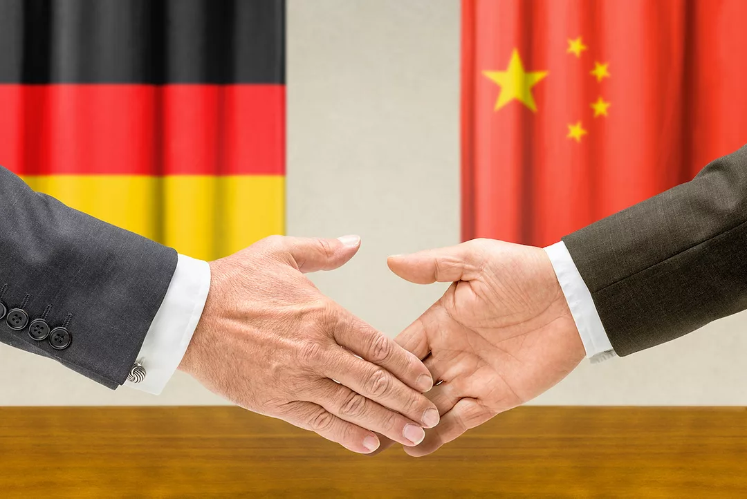 TWO ARMS CAN BE SEEN WITH OPEN HANDS MOVING TOWARDS EACH OTHER FOR A HANDSHAKE WITH THE GERMAN AND CHINESE FLAGS BEHIND EACH ARM IN THE BACKGROUND.