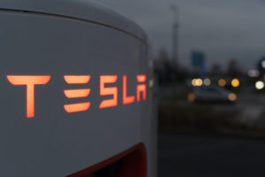 A TESLA SUPERCHARGING STATION CAN BE SEEN AT NIGHT. THE LETTERS SPELLING OUT TESLA ARE ILLUMINATED BY AN ORAGNE BACKLIGHT.