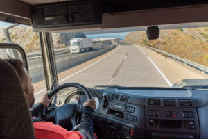 A PERSON IN A RED SHIRT IS DRIVING A TRUCK AND LOOKING OUT THE FRONT WINDOW OUT AT A HIGHWAY. ANOTHER TRUCK IS PASSING BY THE TRUCKER ON THE OTHER SIDE OF THE HIGHWAY.
