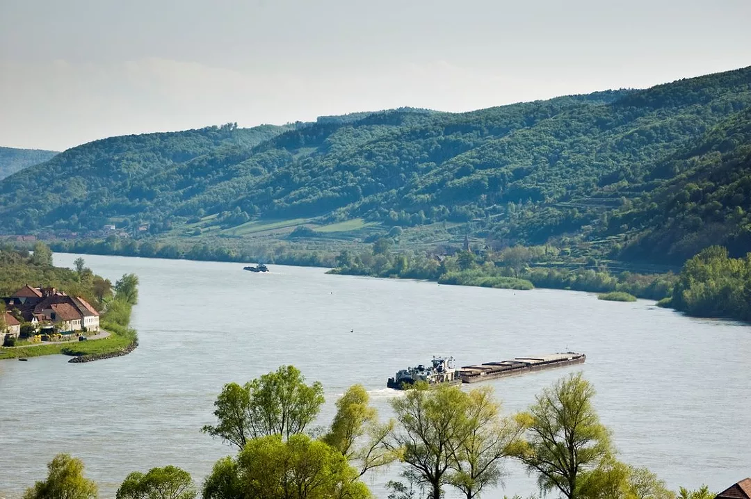 A LOW-LYING CARGO SHIP NAVIGATES A BEND IN A WIDE RIVER, BORDERED BY FORESTS AND A VILLAGE