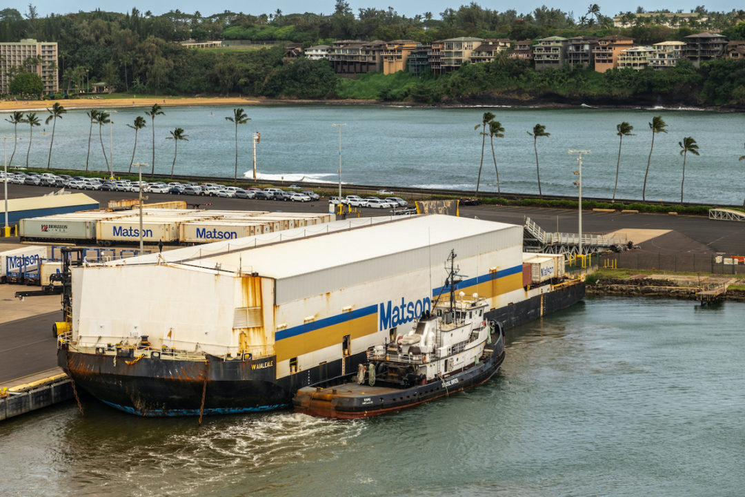 A SMALL BOAT IS DOCKED NEXT TO A WHITE MATSON-LABELED FLOATING BARGE AT A HAWAIIAN PORT.