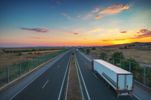 A SEMI-TRAILER TRUCK IS DRIVING BY ITSELF ON A HIGHWAY WHILE THE SUN IS SETTING SURROUNDED BY FIELDS ON BOTH SIDES.