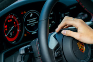 A PERSON'S HAND RESTS ON A STEERING WHEEL THAT SPORTS THE PORSCHE LOGO. THE PORSCHE'S DASHBOARD CAN BE SEEN BEHIND THE WHEEL.