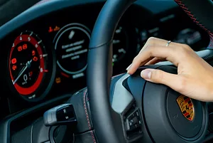 A PERSON'S HAND RESTS ON A STEERING WHEEL THAT SPORTS THE PORSCHE LOGO. THE PORSCHE'S DASHBOARD CAN BE SEEN BEHIND THE WHEEL.