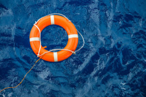AN ORANGE AND WHITE LIFE PRESERVER SITS ON TOP OF A BODY OF WATER WITH THE ROPE DESCENDING OUT OF FRAME. 